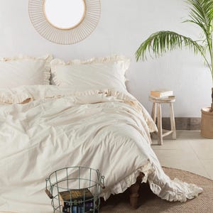 May include: A bedroom scene featuring a bed with cream-colored bedding and ruffled edges. A round mirror hangs on the white wall above the bed. A wire basket filled with books sits on a woven rug. A small wooden stool and a potted plant complete the decor.