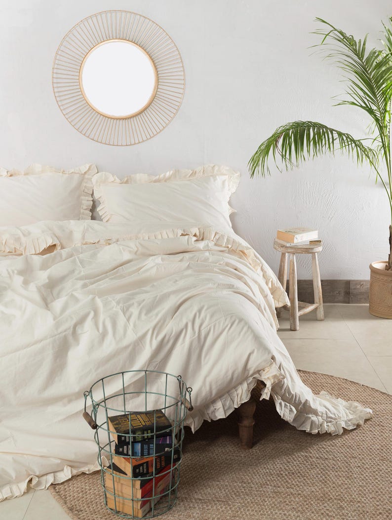 May include: A bedroom scene featuring a bed with cream-colored bedding and ruffled edges. A round mirror hangs on the white wall above the bed. A wire basket filled with books sits on a woven rug. A small wooden stool and a potted plant complete the decor.