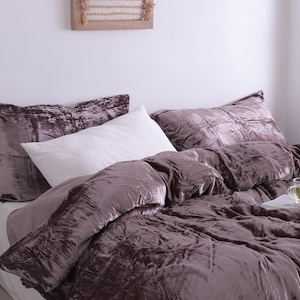 May include: A bed with a plush, velvet-like comforter and matching pillowcases in a muted mauve color. A crisp white pillow provides contrast. The bedding is arranged on a white sheet, creating a cozy and inviting bedroom scene.