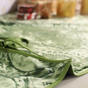 Puede incluir: Primer plano de una falda de &aacute;rbol de Navidad de terciopelo verde con un estampado floral. La falda tiene un borde verde oscuro y un cierre de lazo. El fondo est&aacute; borroso, mostrando regalos envueltos y velas.