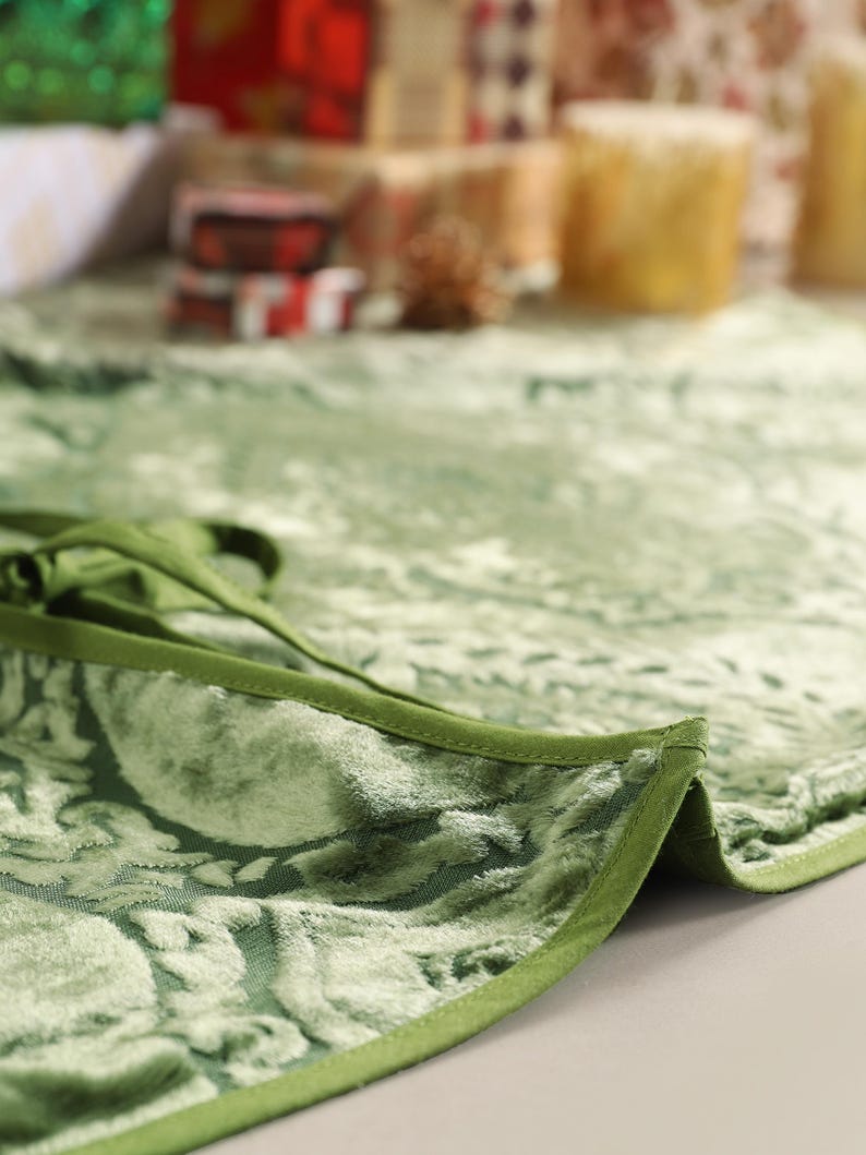 Puede incluir: Primer plano de una falda de &aacute;rbol de Navidad de terciopelo verde con un estampado floral. La falda tiene un borde verde oscuro y un cierre de lazo. El fondo est&aacute; borroso, mostrando regalos envueltos y velas.