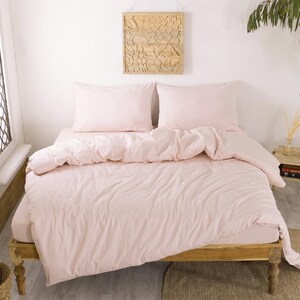 May include: A light pink bed set on a wooden bed frame. The set includes a fitted sheet, duvet cover, and two pillowcases. The bedding is neatly arranged, creating a cozy and inviting atmosphere. The room is bright and airy, with natural light.