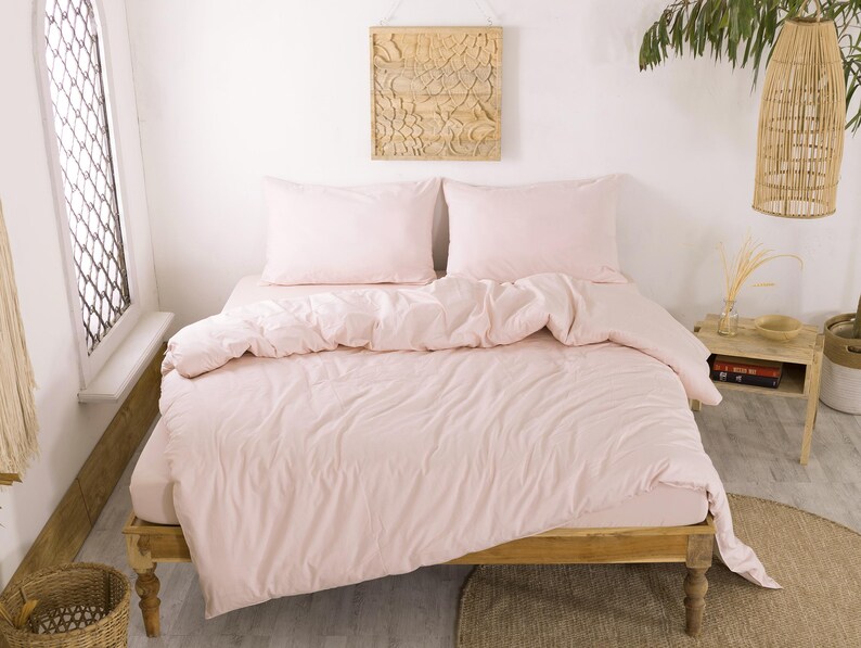 May include: A light pink bed set on a wooden bed frame. The set includes a fitted sheet, duvet cover, and two pillowcases. The bedding is neatly arranged, creating a cozy and inviting atmosphere. The room is bright and airy, with natural light.