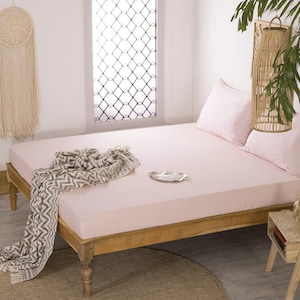 Puede incluir: Una cama rosa claro con almohadas a juego y un marco de madera. Una manta con estampado gris y blanco está sobre la cama. Un atrapasueños tejido y una lámpara decorativa cuelgan cerca. Una pequeña mesita de noche de madera está al lado de la cama.