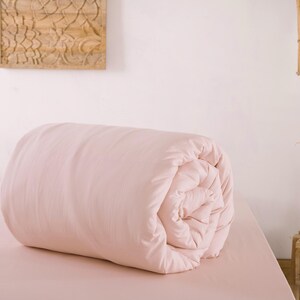 May include: A rolled-up, light pink comforter rests on a matching bed. The bedding is the focal point, with a soft, inviting appearance. The scene is set in a bedroom, with a wooden wall decoration and a small side table visible.