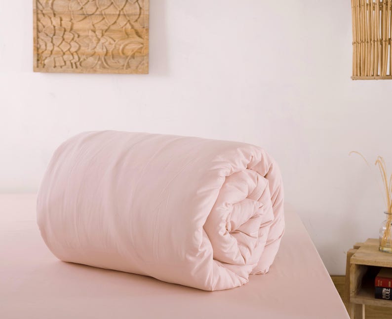 May include: A rolled-up, light pink comforter rests on a matching bed. The bedding is the focal point, with a soft, inviting appearance. The scene is set in a bedroom, with a wooden wall decoration and a small side table visible.