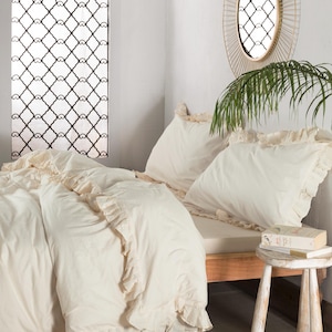 May include: Cream-colored bedding set with ruffled edges. The set includes a duvet cover and pillowcases, arranged on a wooden bed frame. A small wooden stool holds books. A decorative window and mirror are visible in the background.