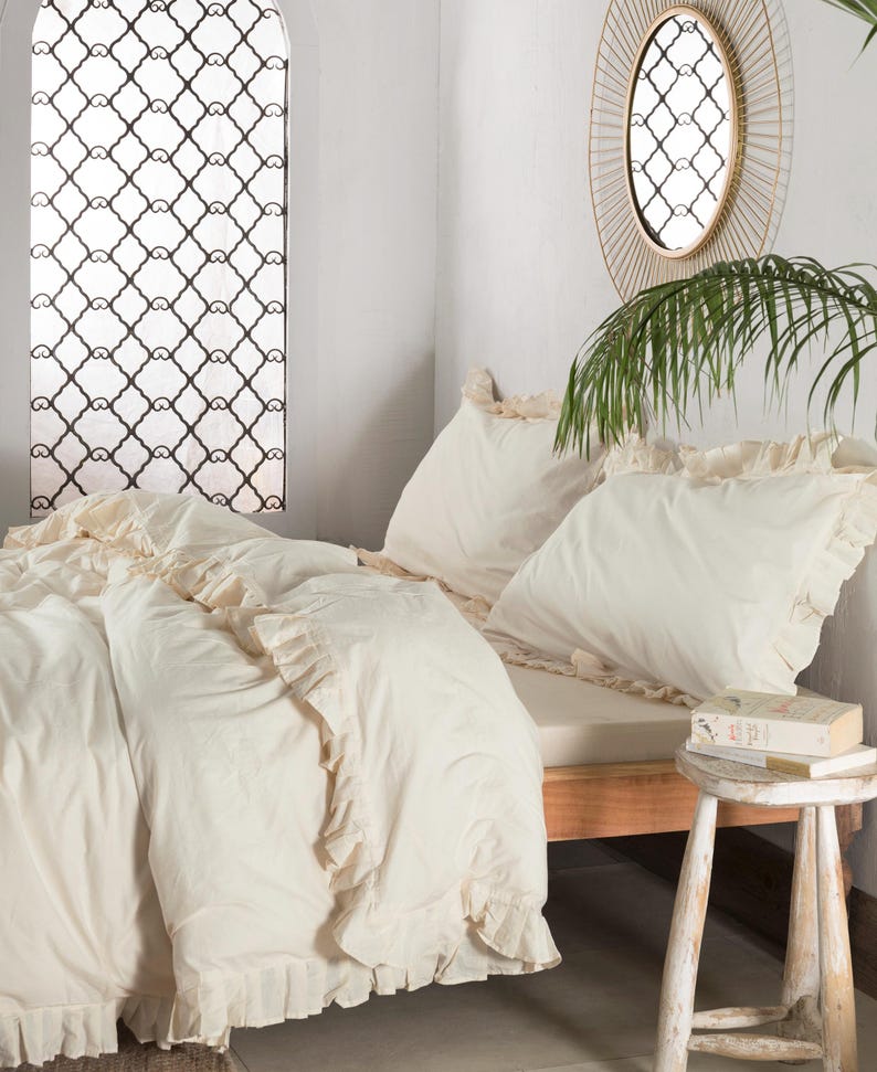 May include: Cream-colored bedding set with ruffled edges. The set includes a duvet cover and pillowcases, arranged on a wooden bed frame. A small wooden stool holds books. A decorative window and mirror are visible in the background.