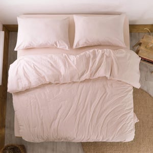 May include: Overhead view of a bed with light pink bedding. Two matching pillowcases sit atop the bed, with a comforter and fitted sheet in the same soft pink hue. The bed is set against a light-colored wall and floor.