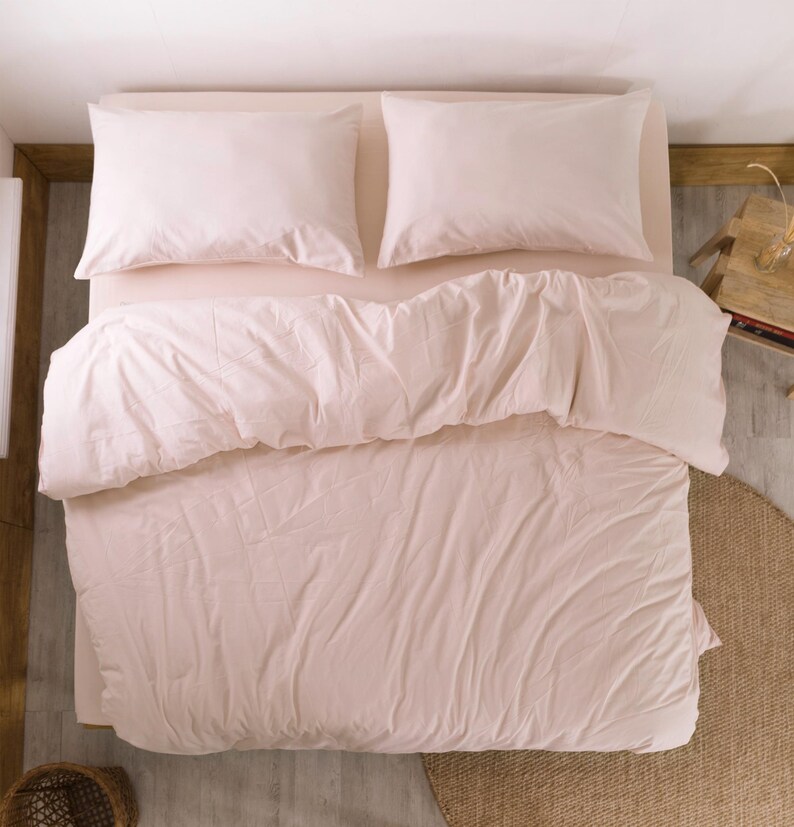 May include: Overhead view of a bed with light pink bedding. Two matching pillowcases sit atop the bed, with a comforter and fitted sheet in the same soft pink hue. The bed is set against a light-colored wall and floor.