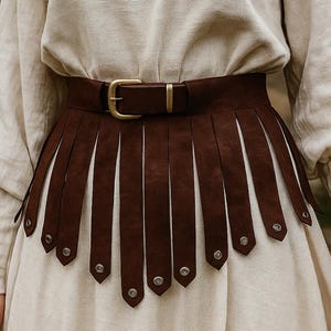 May include: A brown leather belt with a decorative skirt-like design. The belt has a gold-colored buckle and is worn over a cream-colored dress. The skirt-like design has vertical strips and silver rivets.
