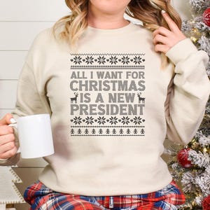 Può includere: Una felpa color crema con la scritta "ALL I WANT FOR CHRISTMAS IS A NEW PRESIDENT" in nero. Il design include motivi a fiocco di neve e renne. Una tazza bianca è tenuta in mano. Abbigliamento festivo per le festività.