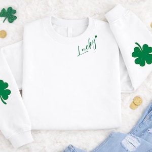 Può includere: Felpa bianca con dettagli verdi, con un quadrifoglio sulla manica e la scritta "Lucky" in verde. Monete d'oro e decorazioni a trifoglio sono sparse intorno al capo, suggerendo un tema del giorno di San Patrizio.