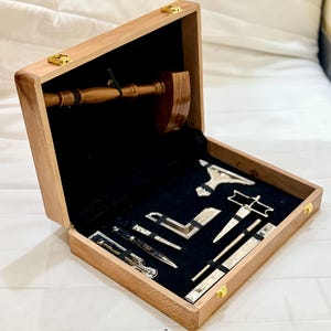 May include: A wooden box with a black velvet interior, housing a gavel and several silver tools. The box is crafted from light brown wood and features gold-coloured clasps.