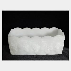 May include: A white rectangular dish with a scalloped rim. The sides of the dish are decorated with a grape and vine pattern. The dish is made of a solid white material.