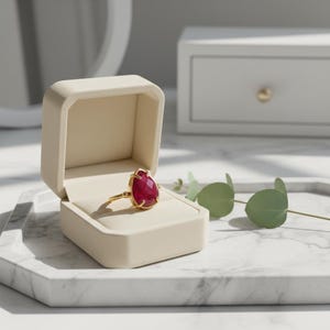 May include: A gold ring with a large, teardrop-shaped, red gemstone sits in an open, beige ring box. The ring box is on a marble tray with a sprig of green leaves. A white chest of drawers and mirror are in the background.