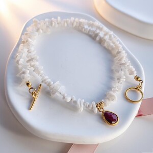 May include: A white gemstone necklace with a gold-coloured clasp and a teardrop-shaped red pendant. The necklace is made of small, irregular white stones. The pendant is set in a gold-coloured frame. The necklace is displayed on a white surface.