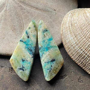 May include: Two polished gemstone pieces with a triangular shape, featuring a blend of light green, turquoise, and black colors. The stones are set against a backdrop of a sand dollar and a seashell.