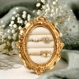 Peut inclure: Un présentoir à bijoux ovale doré orné contenant quatre bagues et une paire de boucles d'oreilles. Les bagues comprennent une bague de perle, une bague sertie de diamants et une bague avec de petites pierres. Les boucles d'oreilles sont des boucles d'oreilles nœud perle.