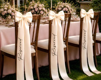 May include: Three wooden chairs with white upholstered seats, each featuring a large cream-colored satin ribbon tied into a bow on the backrest. The long ribbon tails hang down and are printed with the word Reserved in a black cursive script.