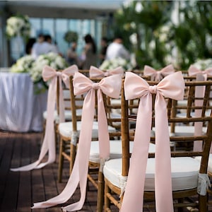 Peut inclure: Rangée de chaises en bois avec coussins blancs, ornées de grands nœuds en satin rose pâle et de rubans flottants. Les chaises sont disposées pour un événement en plein air, avec des silhouettes floues et des compositions florales en arrière-plan.