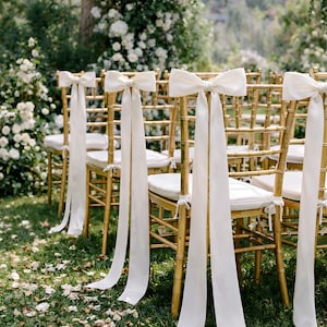 Pode incluir: Cadeiras douradas de bambu com almofadas brancas e grandes laços brancos amarrados nas costas, dispostas em um gramado. O fundo apresenta flores brancas e vegetação, sugerindo um casamento ou evento especial.