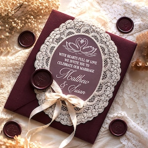 May include: A burgundy wedding invitation with a white lace oval frame and a cream ribbon. The invitation includes the text "With hearts full of love, we invite you to celebrate our marriage, Matthew & Susan." Burgundy wax seals are scattered around the invitation.