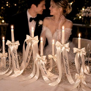 May include: A set of clear glass candle holders with white taper candles, each adorned with cream-coloured satin ribbons tied in bows. The ribbons feature the printed text "Just Married" and the date "04-10-2026" in a black script font.