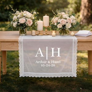 Peut inclure: Une table en bois est dressée pour une réception de mariage, avec un chemin de table blanc transparent orné d'une délicate bordure en dentelle. Le chemin de table affiche les initiales "A H", les noms "Arthur & Hazel" et la date "05-24-26". Des compositions florales et des bougies complètent le décor élégant.