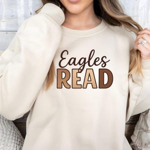 May include: Cream-colored sweatshirt with the words "Eagles READ" in brown and tan lettering. The sweatshirt is a neutral color and has a crew neck. The text is in a playful, readable font.
