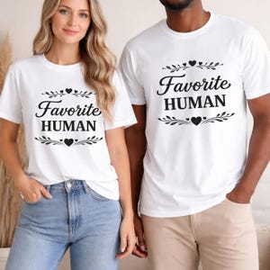 May include: Two people wearing white t-shirts with the text "Favorite HUMAN" in black script. The design includes decorative leaves and hearts. The shirts are short-sleeved and the people are wearing blue jeans and khaki pants.