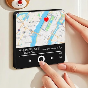 May include: A black rectangular magnet with a map of New York City, a red heart, and the text "WHERE WE MET". The magnet has a music player interface with play, pause, and skip buttons. The magnet is being held by a person.