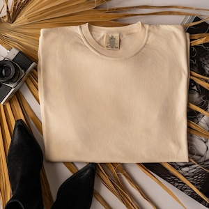 Op de afbeelding: Een gevouwen, crèmekleurig t-shirt met een ronde hals, liggend op een wit oppervlak. Een vintage camera, zwarte laarzen en een gouden ketting zijn rond het shirt gerangschikt. Gedroogde palmbladeren voegen een natuurlijk element toe.