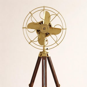 Può includere: Un ventilatore da terra in stile retrò, con gabbia e pale in ottone, montato su un treppiede in legno scuro. Il ventilatore riporta la scritta "Made for Royal Navy London". La base del treppiede presenta dettagli in ottone e un cavo di alimentazione nero.