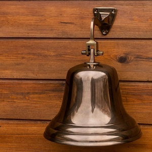 May include: A shiny brass bell fixed to a wooden wall. The bell has a wide base and a curved arm connected to a triangular mounting plate. The wood has a warm brown colour with visible grain.