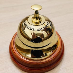 May include: A shiny gold-colored bell with a wooden base. The bell has the text "HUGGING BELL RING FOR A BIT" engraved on its surface. The bell is designed with a spring-loaded clapper and a small knob on top.