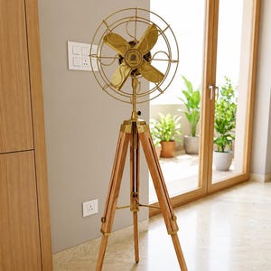 May include: A floor-standing fan featuring a brass head with four blades and a protective wire cage, mounted on a tripod base made of light-toned wood with brass hardware accents. The centre of the fan head displays a circular emblem with text.