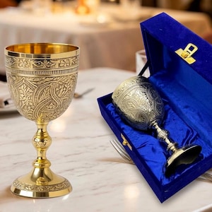 May include: Ornate gold-colored chalice with intricate floral and leaf patterns. The chalice is displayed on a marble surface, with a second chalice inside a blue velvet presentation box. The chalice has a detailed base and stem.