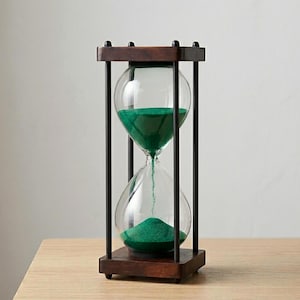 May include: An hourglass featuring a dark brown wooden frame with four vertical black metal rods connecting the top and bottom square bases. The clear glass bulb contains fine green sand, with a portion currently flowing through the center.