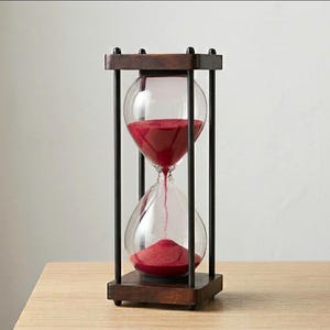 Könnte beinhalten: Eine Sanduhr mit einem dunkelbraunen Holzrahmen und vier schwarzen Metallstützstangen. Der klare Glaskolben enthält leuchtend roten Sand, wobei ein Teil des Sandes gerade durch die schmale Mitte in die untere Kammer fließt.