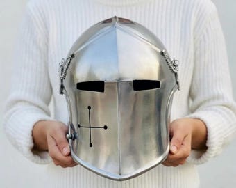 May include: A polished silver knight's helmet with a cross design on the front. The helmet has eye slits and is held up by a person wearing a white sweater. The helmet has chain details on the sides.