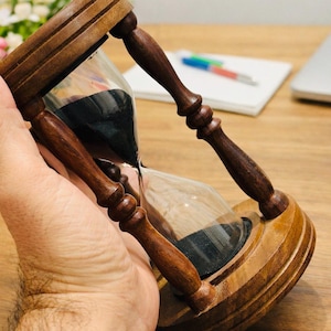 以下が含まれることがあります： 手で持たれた、内部に暗い砂が入った木製の砂時計。砂時計は、細い首でつながれた、膨らんだ上部と下部を備えたクラシックなデザインです。木製のフレームは濃い茶色です。背景には、ノート、ペン、ラップトップが含まれています。