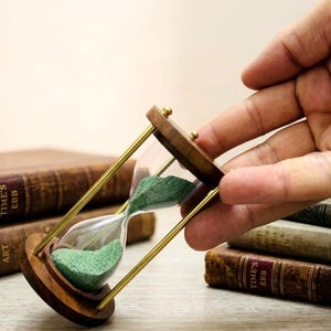 Op de afbeelding: Een zandloper met een houten frame en messing steunen, gevuld met groen zand. De zandloper wordt vastgehouden in de buurt van een stapel antieke boeken met versleten, bruine omslagen. De boeken hebben gouden letters.