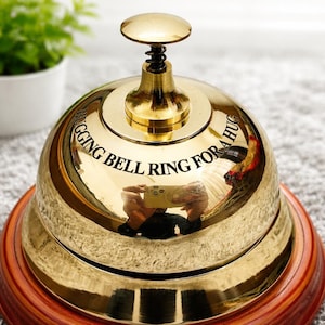 Peut inclure: Une cloche dorée avec une base en bois. L'inscription "HUGGING BELL RING FOR A HUG" est gravée sur la cloche. Un petit pot blanc avec une plante verte est visible en arrière-plan.