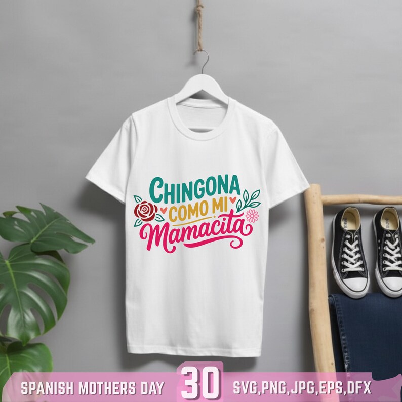 May include: White t-shirt with the text "Chingona Como Mi Mamacita" in teal and pink, with floral accents. The shirt is hanging on a white hanger. The words "Spanish Mothers Day" and the number "30" are at the bottom.