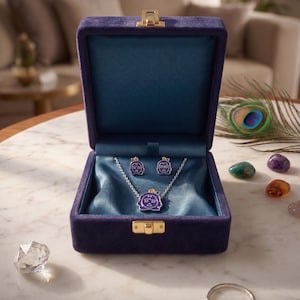Peut inclure: Un ensemble de bijoux composé d'un collier à chaîne argentée et de boucles d'oreilles assorties, chacune dotée d'un pendentif en émail violet avec un motif de visage stylisé. L'ensemble est présenté dans un coffret doublé de velours violet.