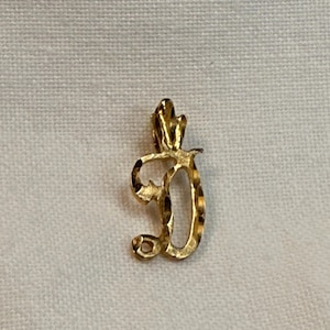 May include: A gold-colored pendant in the shape of the letter "D". The pendant has a textured surface and a small loop at the top for a chain. The design is ornate and elegant, suitable for jewelry.