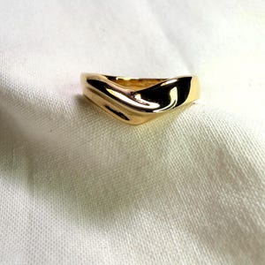 May include: A polished gold ring featuring a smooth, continuous band with a distinct V-shaped chevron contour. The metal surface is highly reflective, showcasing a curved, fluid design that tapers slightly toward the back of the band.