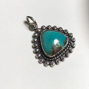 May include: A triangular turquoise pendant set in a silver frame. The stone features a vibrant blue-green color with dark brown matrix inclusions. The silver setting includes a twisted rope border surrounded by a row of small silver beads.