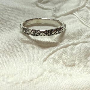 May include: A silver-toned metal ring featuring a continuous band with a repeating engraved pattern of small diamond shapes and vertical lines. The surface of the ring has a polished finish with darkened recessed areas that highlight the intricate design.